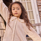 A young girl with long wavy hair poses outdoors, one arm raised, wearing the Bonton Girls’ Figue Shirt—a cream 100% cotton button-up featuring delicate lace trim and a charming Peter Pan collar.