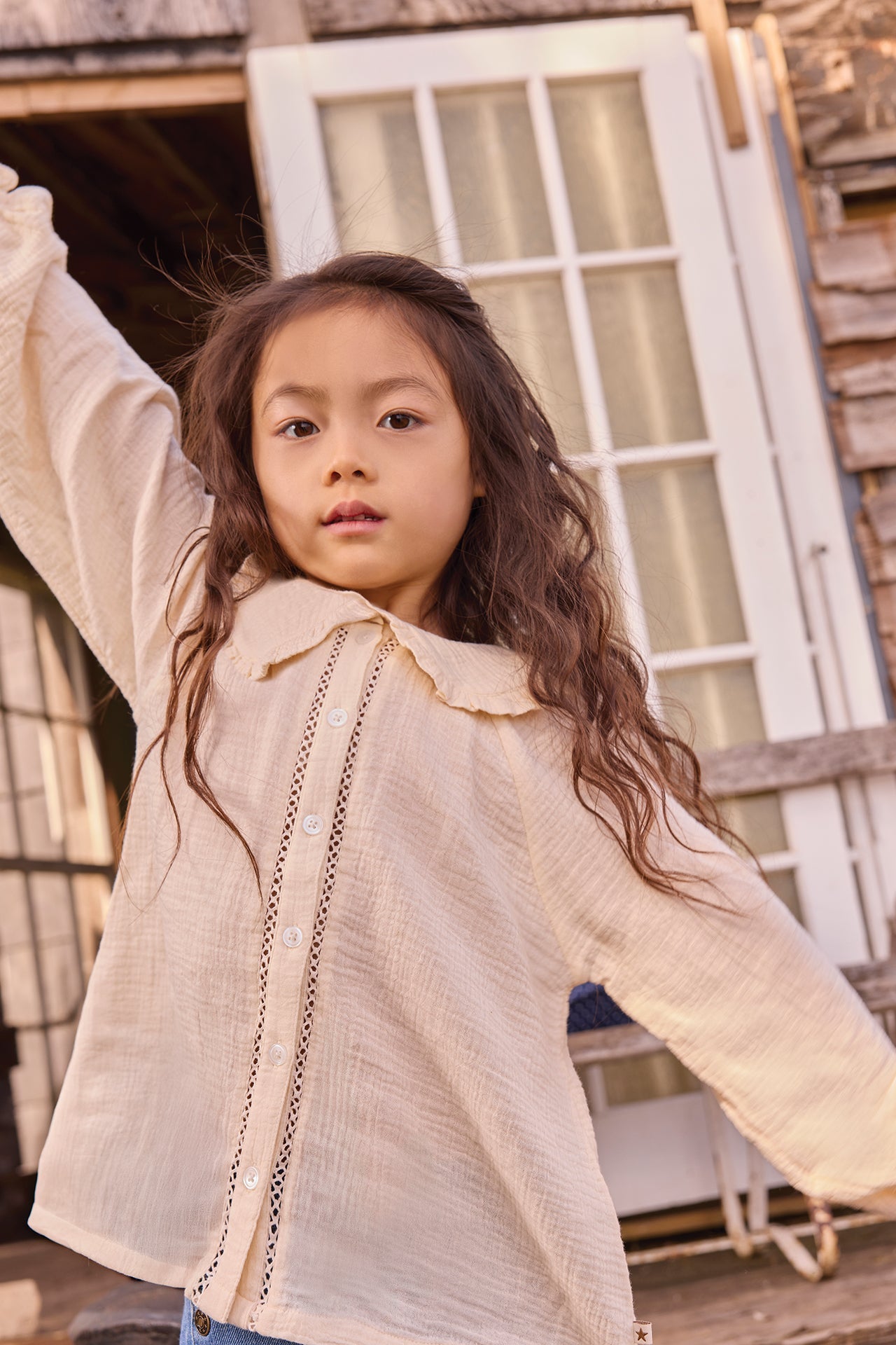 A young girl with long wavy hair poses outdoors, one arm raised, wearing the Bonton Girls’ Figue Shirt—a cream 100% cotton button-up featuring delicate lace trim and a charming Peter Pan collar.