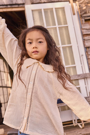 A young girl with long wavy hair poses outdoors, one arm raised, wearing the Bonton Girls’ Figue Shirt—a cream 100% cotton button-up featuring delicate lace trim and a charming Peter Pan collar.