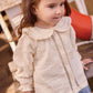 A young child with long brown hair stands outdoors, wearing a cream-colored Bonton Baby Girls’ and Toddler Fig Shirt with ruffles and blue pants.