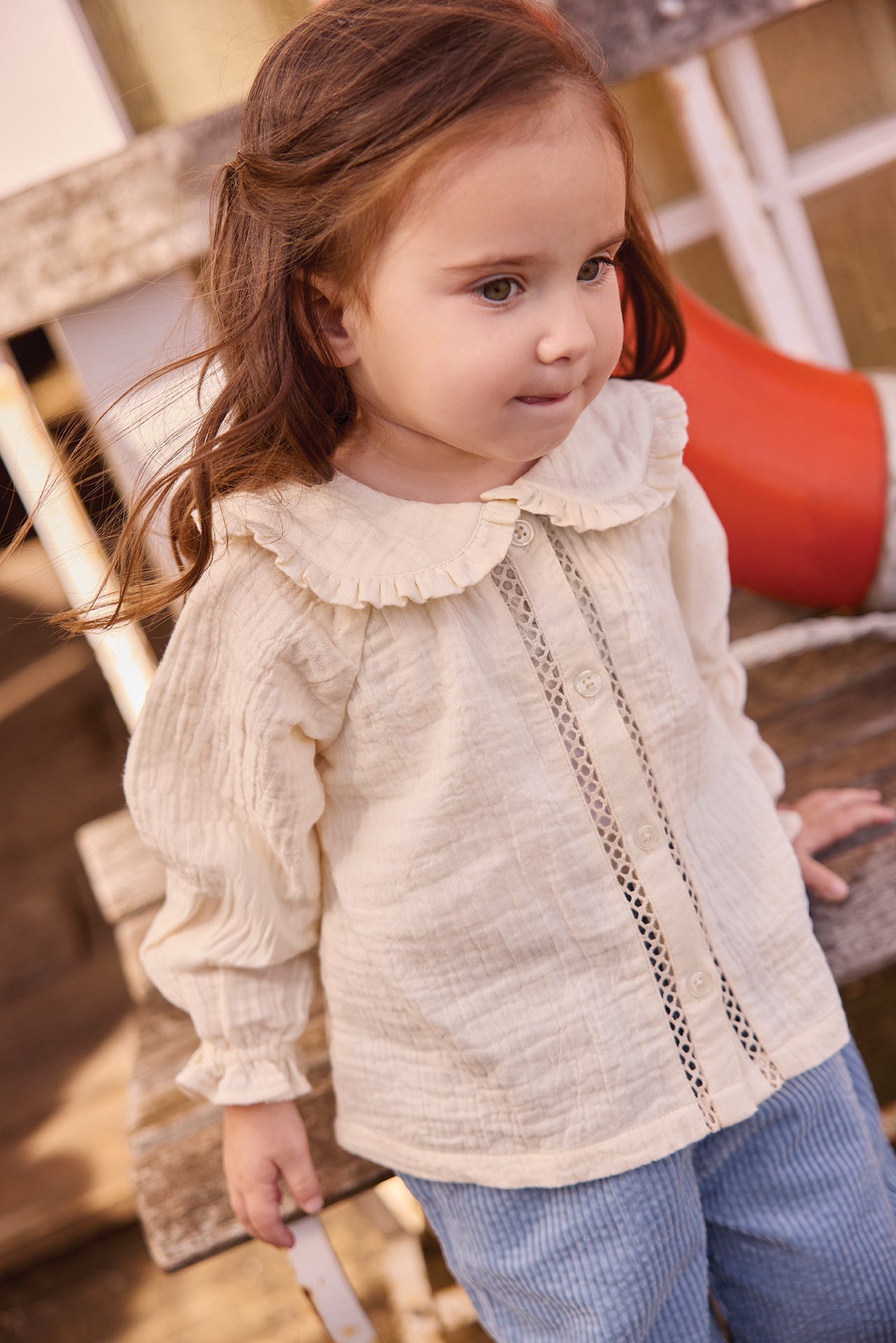 A young child with long brown hair stands outdoors, wearing a cream-colored Bonton Baby Girls’ and Toddler Fig Shirt with ruffles and blue pants.