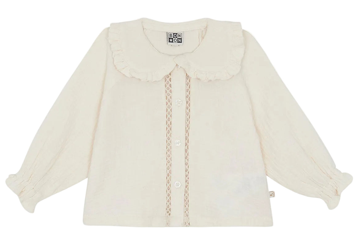 The Baby Girls’ and Toddler Fig Shirt by Bonton is a cream-colored long-sleeve shirt featuring a ruffled Peter Pan collar, front buttons, lace trim, and elastic cuffs, displayed on a white background.
