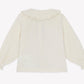 Back view of the Bonton Baby Girls’ and Toddler Fig Shirt, a long-sleeved, off-white top featuring a Peter Pan collar with delicate ruffles and gathered cuffs, displayed on a plain background.