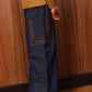 A child is wearing the Bonton Girls’ Lauren Pant—crafted from comfortable chambray cotton for all-day ease—in dark blue with wide legs and a large decorative pocket, paired with a brown sweater featuring white hearts and silver shoes, standing against a wood-paneled wall and parquet floor.