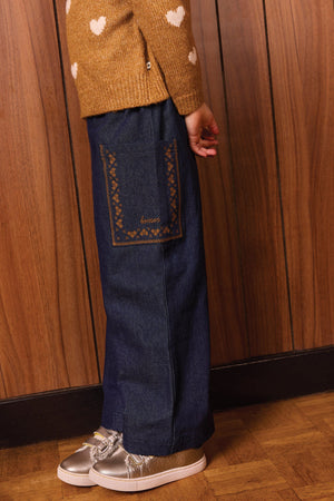 A child is wearing the Bonton Girls’ Lauren Pant—crafted from comfortable chambray cotton for all-day ease—in dark blue with wide legs and a large decorative pocket, paired with a brown sweater featuring white hearts and silver shoes, standing against a wood-paneled wall and parquet floor.