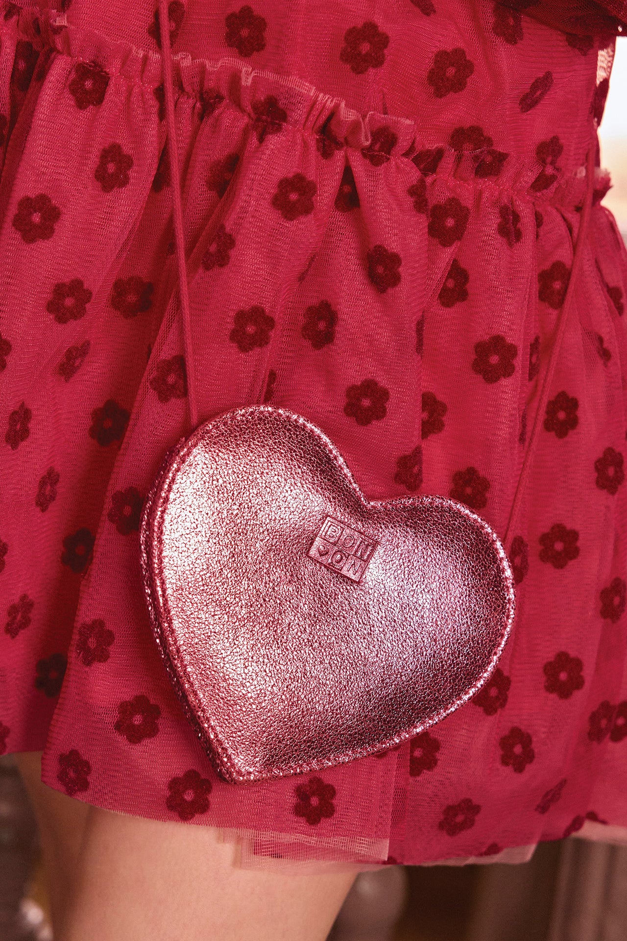 Close-up of a person wearing a red skirt with floral patterns, carrying the Bonton Girls’ Leather Heart Bag in shiny pink leather with a small embossed logo.