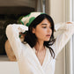 A woman with long dark hair adjusts the Bembien Delfina Scarf, a green and white floral design handprinted in Italy, on her head. She wears a white blouse while standing near a window bathed in soft natural lighting.