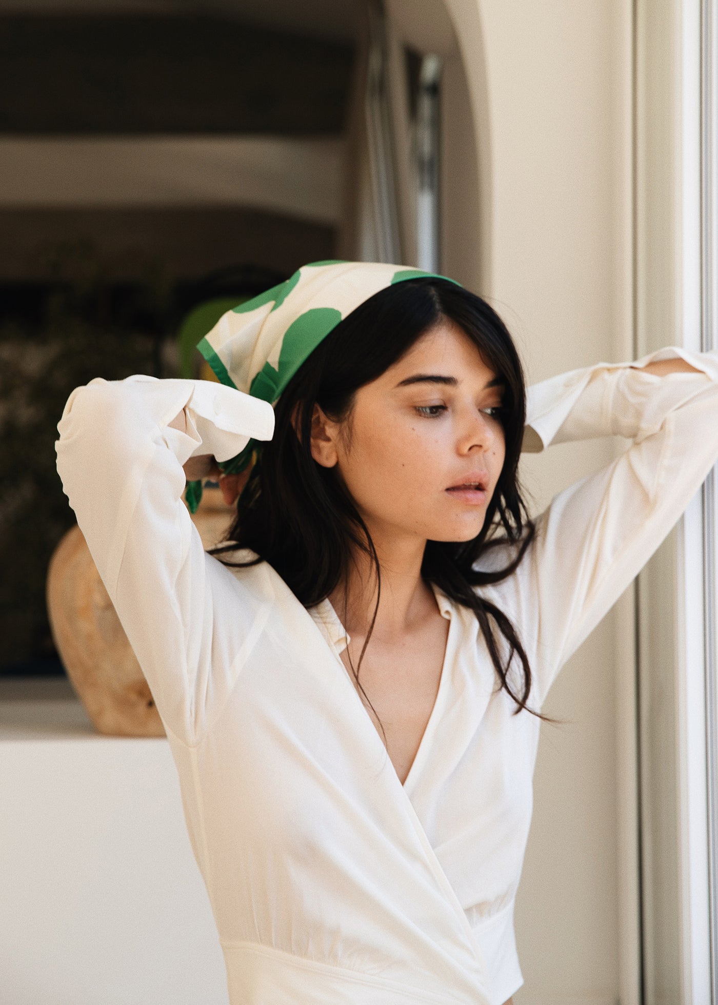 A woman with long dark hair adjusts the Bembien Delfina Scarf, a green and white floral design handprinted in Italy, on her head. She wears a white blouse while standing near a window bathed in soft natural lighting.