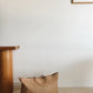 The Bembien Le Traveler, a large woven cow leather tote bag with an adjustable strap, is placed on the floor next to a wooden table against a white wall. A framed abstract print is hung on the wall.