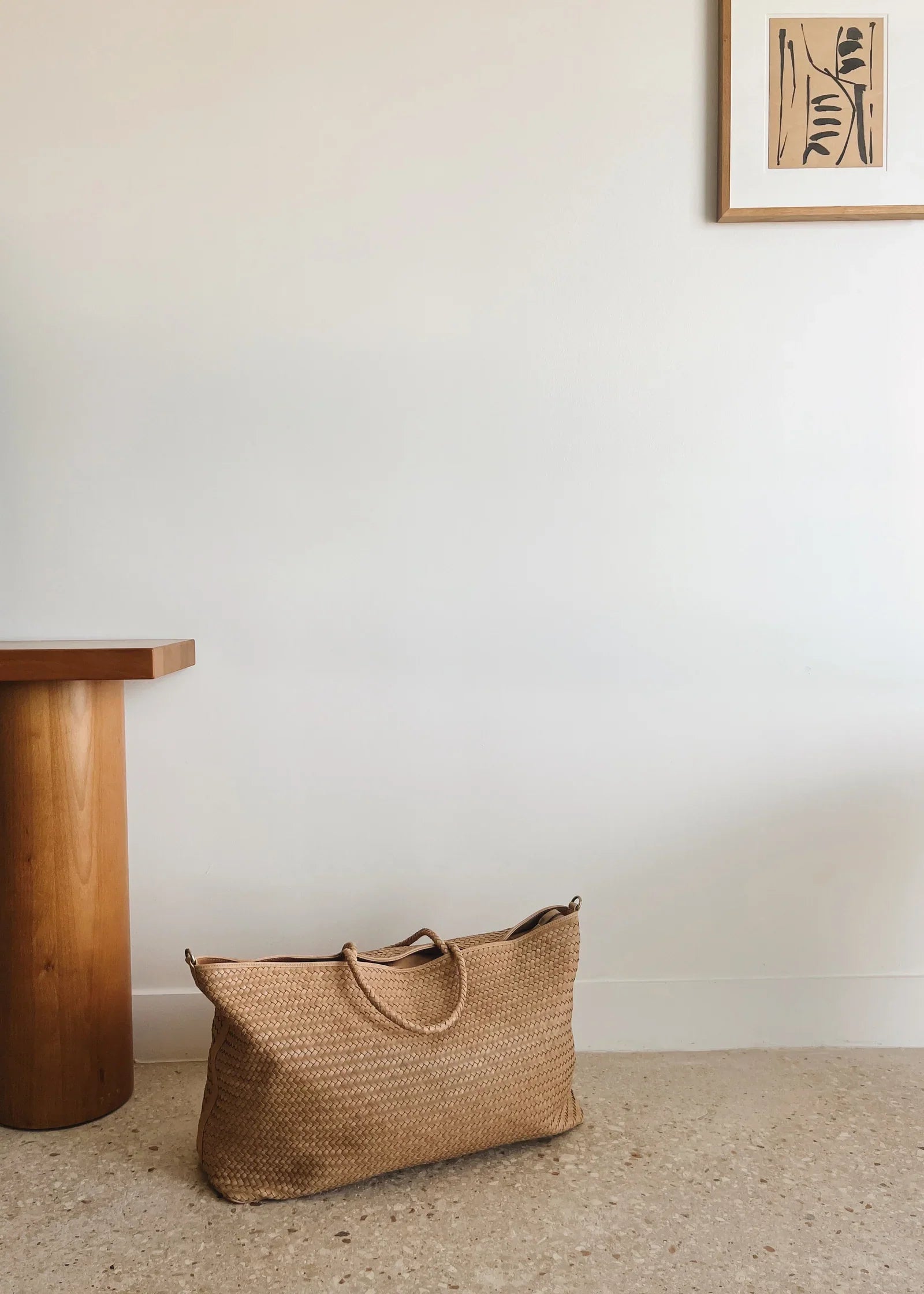 The Bembien Le Traveler, a large woven cow leather tote bag with an adjustable strap, is placed on the floor next to a wooden table against a white wall. A framed abstract print is hung on the wall.