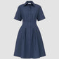 The Bergamo Dress by Callas Milano is a navy blue, short-sleeve, collared, button-down fit and flare cotton poplin dress with a pleated front and fitted waist, shown against a plain light gray background.
