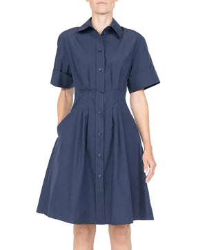 A person wears the Callas Milano Bergamo Dress—a navy blue, short-sleeve, button-front fit-and-flare dress with pleats and side pockets in cotton poplin—while standing against a plain background.