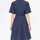 A person stands facing away, wearing the Callas Milano Bergamo Dress—a short-sleeved, dark blue cotton poplin fit and flare style—against a plain white background.