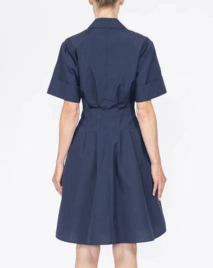 A person stands facing away, wearing the Callas Milano Bergamo Dress—a short-sleeved, dark blue cotton poplin fit and flare style—against a plain white background.