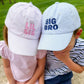 Two kids sit outdoors in green grass, both wearing Bits & Bows caps—her pink hat and his blue "Big Bro Baseball Hat." The siblings relax together, styled in classic headwear that captures true coastal charm.