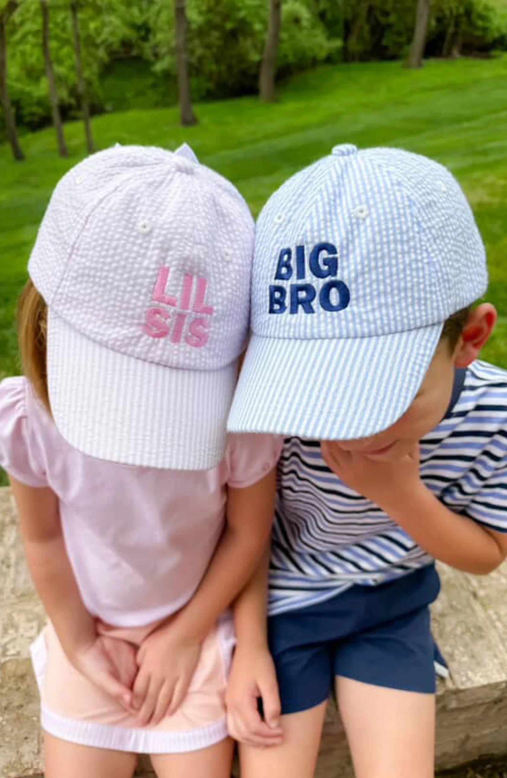 Two kids sit outdoors in green grass, both wearing Bits & Bows caps—her pink hat and his blue "Big Bro Baseball Hat." The siblings relax together, styled in classic headwear that captures true coastal charm.