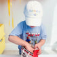 A young child in a Bits & Bows "Birthday Boy Baseball Hat" peers into a red gift bag, surrounded by colorful balloons that add a touch of coastal charm.