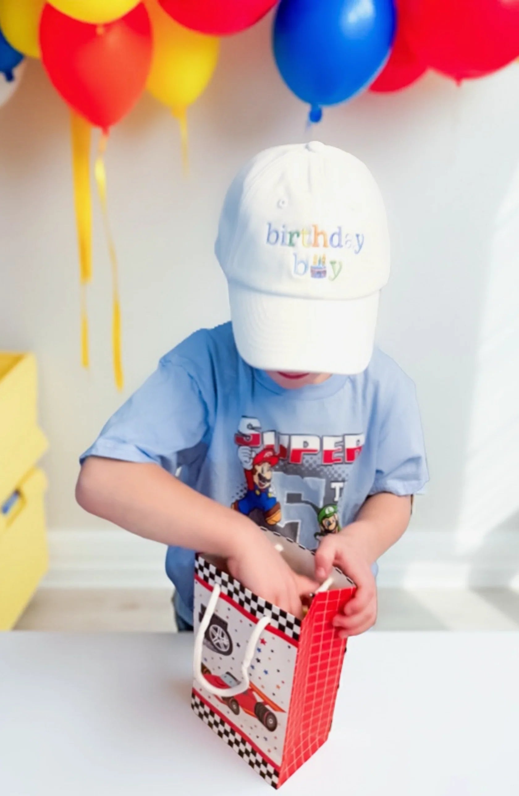 A young child in a Bits & Bows "Birthday Boy Baseball Hat" peers into a red gift bag, surrounded by colorful balloons that add a touch of coastal charm.