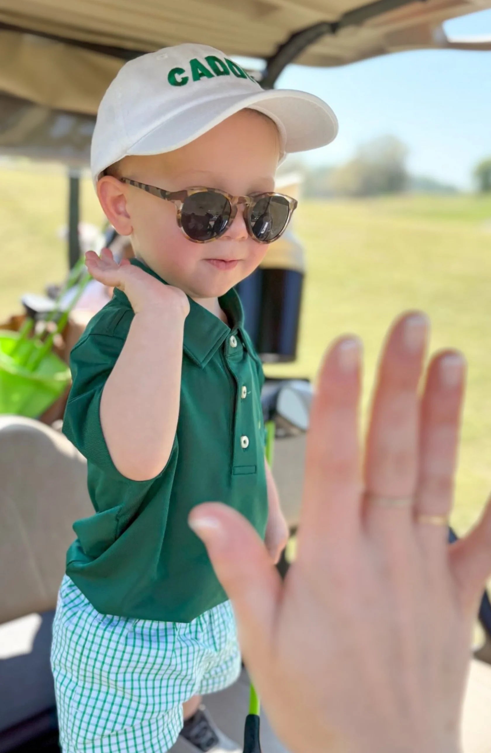 A young child in sunglasses, a green shirt, and a Bits & Bows Caddie Baseball Hat gets ready to high five an outstretched hand outside on a sunny day.