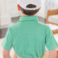 A young boy stands indoors with his back to the camera, wearing the Bits & Bows Caddie Baseball Hat, a green and white striped polo, and beige pants.