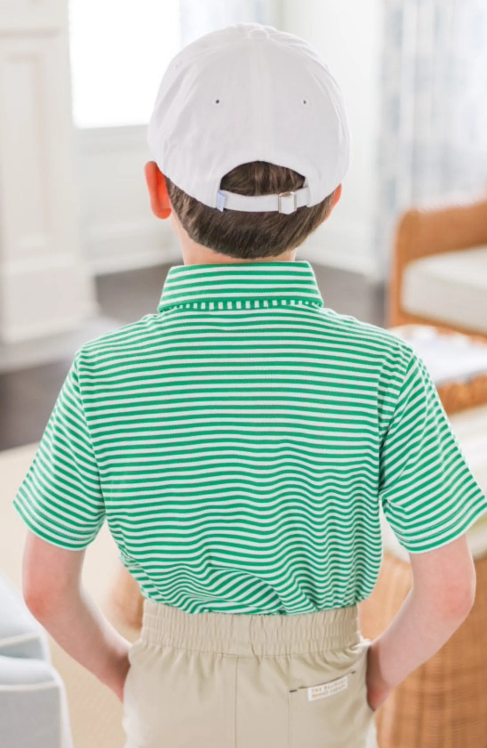 A young boy stands indoors with his back to the camera, wearing the Bits & Bows Caddie Baseball Hat, a green and white striped polo, and beige pants.
