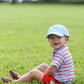 A young child in a Bits & Bows Golf Cart Baseball Hat, striped shirt, and red shorts sits on green grass, smiling at the camera.