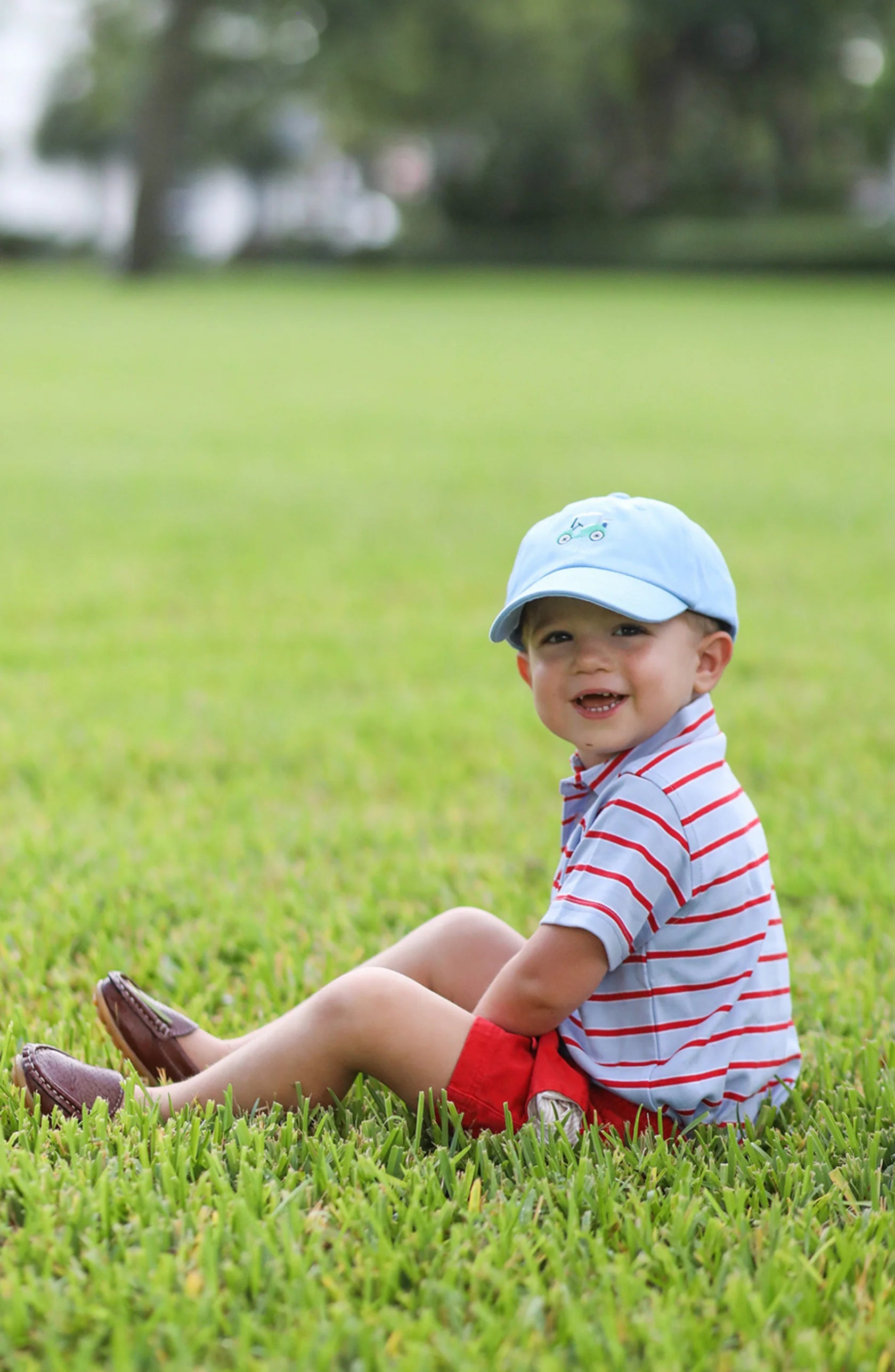 A young child in a Bits & Bows Golf Cart Baseball Hat, striped shirt, and red shorts sits on green grass, smiling at the camera.