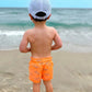 A young child in orange shark-print shorts and a Bits & Bows Blue Seersucker Football Baseball Hat stands on the sandy beach, facing the ocean.