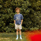 A young boy stands on grass near lush bushes, wearing a blue "Ride the Vibe" shirt, Gingersnaps Boys' Blaine Shorts with Braided Belt, and light blue socks. A red blur is visible in the lower right foreground.