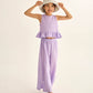 A young girl smiles in the Cozmo Girls' Blair Top—a lilac cotton gauze sleeveless top with a ruffled peplum hem—paired with matching wide-leg pants, a floral hat, and beige shoes against a plain background.