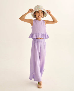 A young girl smiles in the Cozmo Girls' Blair Top—a lilac cotton gauze sleeveless top with a ruffled peplum hem—paired with matching wide-leg pants, a floral hat, and beige shoes against a plain background.