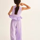 A young girl with long brown hair in a ponytail is seen from behind wearing the Cozmo Girls' Blair Top, a sleeveless lavender peplum style, paired with matching wide-leg pants.