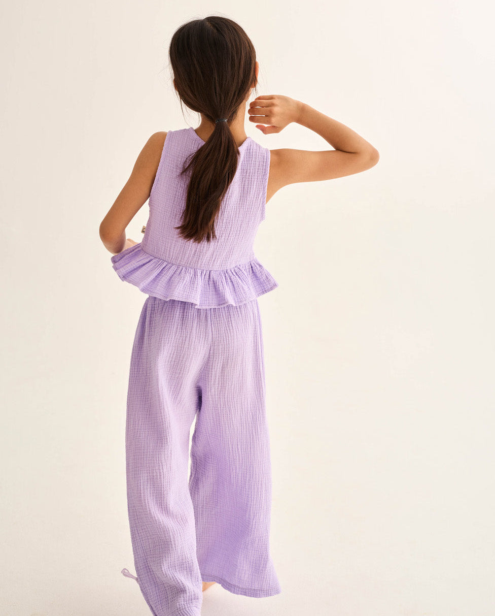 A young girl with long brown hair in a ponytail is seen from behind wearing the Cozmo Girls' Blair Top, a sleeveless lavender peplum style, paired with matching wide-leg pants.