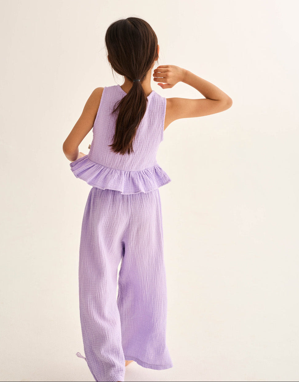 A girl with long brown hair in a ponytail wears Cozmo's Girls' Violet Wide Pants, featuring an elastic waistband, as she stands with her back to the camera against a plain background.