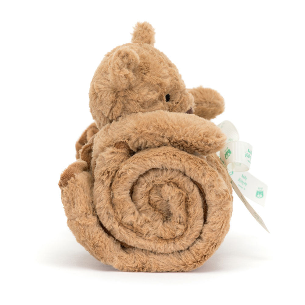 A small, brown Jellycat Bartholomew Bear Blankie teddy bear holding a rolled-up beige blanket tied with a white ribbon featuring green owl prints from the Jellycat series.