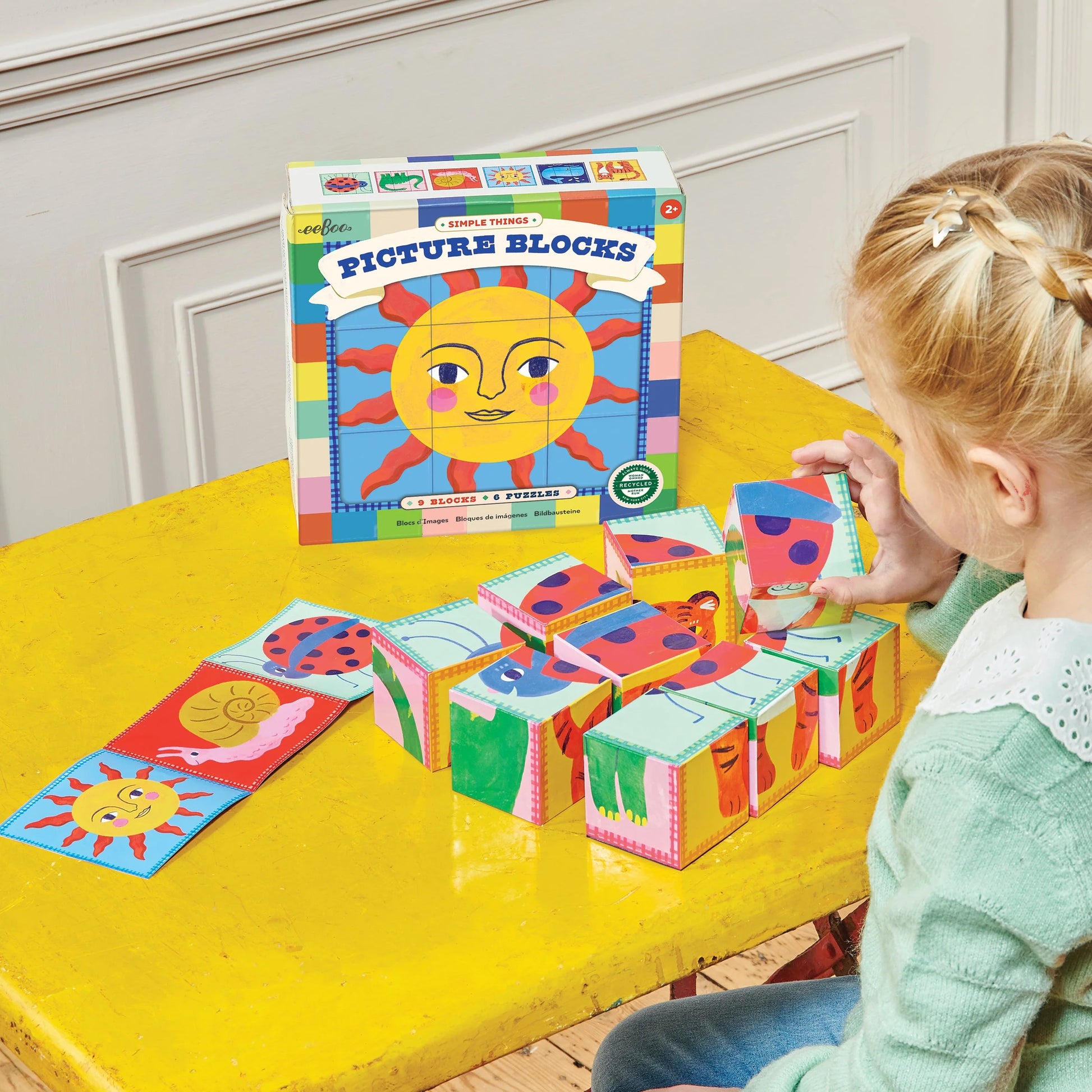 A young girl arranges Eeboo Simple Things Picture Blocks from a 9-block set on a yellow table, with the "Simple Things Picture Blocks" box and completed puzzles displayed beside her.