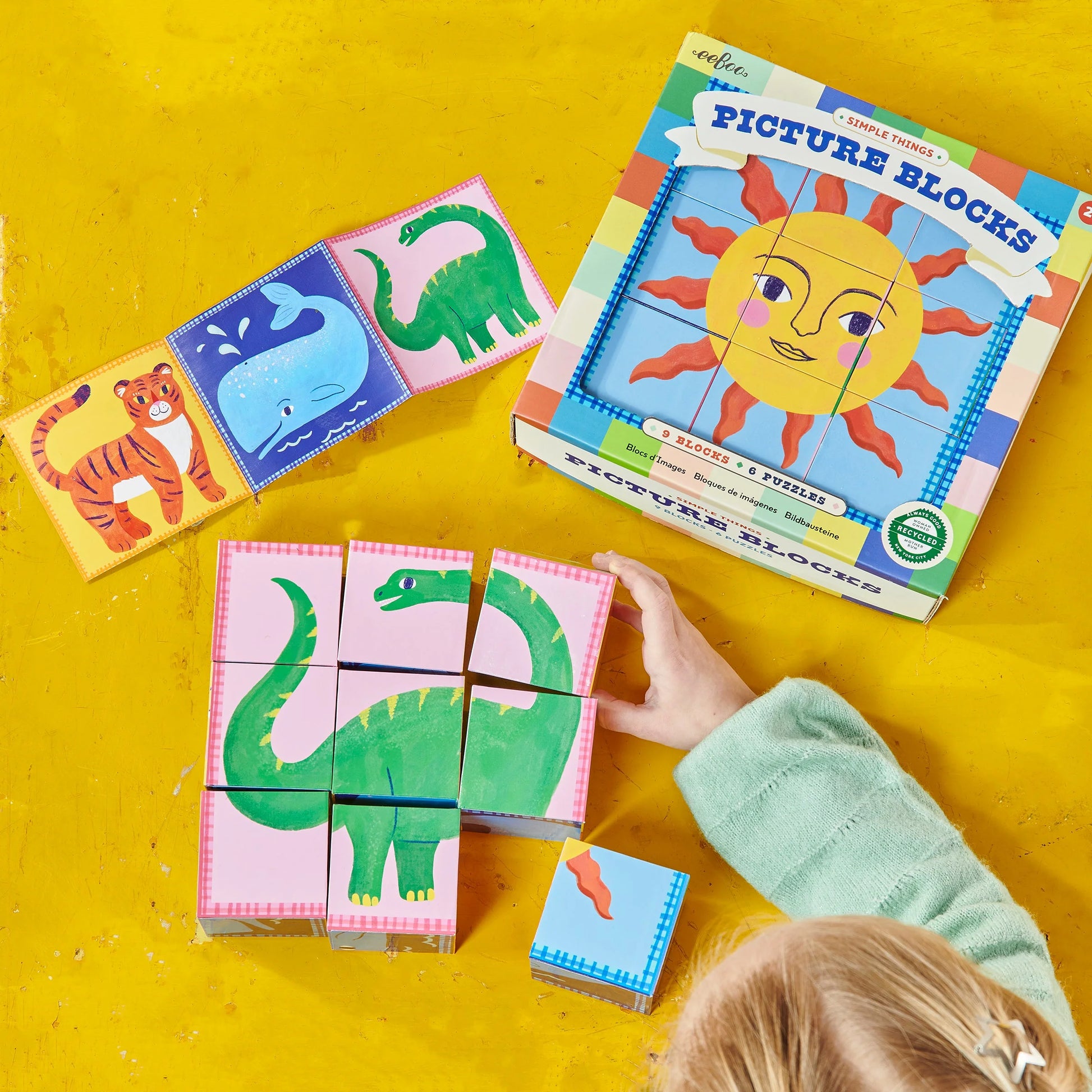 A child arranges Eeboo’s Simple Things Picture Blocks—a 9-block wooden set forming a dinosaur—while the box and easy tiger and whale puzzles sit on a yellow surface.