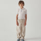 A young boy with curly hair stands against a white background, wearing a short-sleeved white polo shirt, Gingersnaps Boys' Erwin Drawstring Trousers, and light blue sandals.