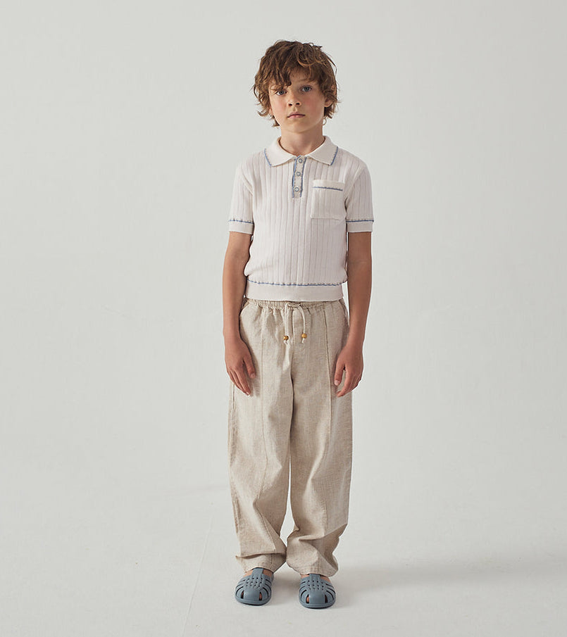 A young boy with curly hair stands against a white background, wearing a short-sleeved white polo shirt, Gingersnaps Boys' Erwin Drawstring Trousers, and light blue sandals.