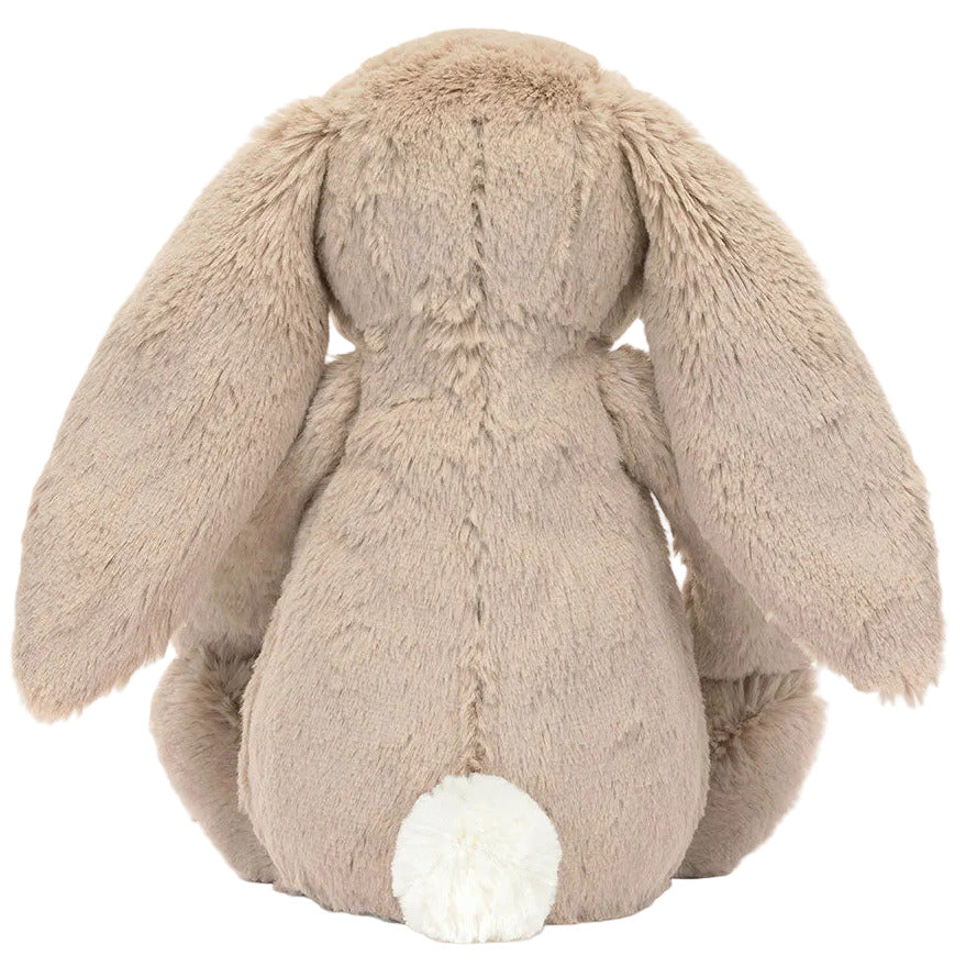 The Jellycat Blossom Beige Bunny 'Petal' sits facing away, displaying its long floppy ears and tiny white tail, as if lost in a daydream among wildflowers.