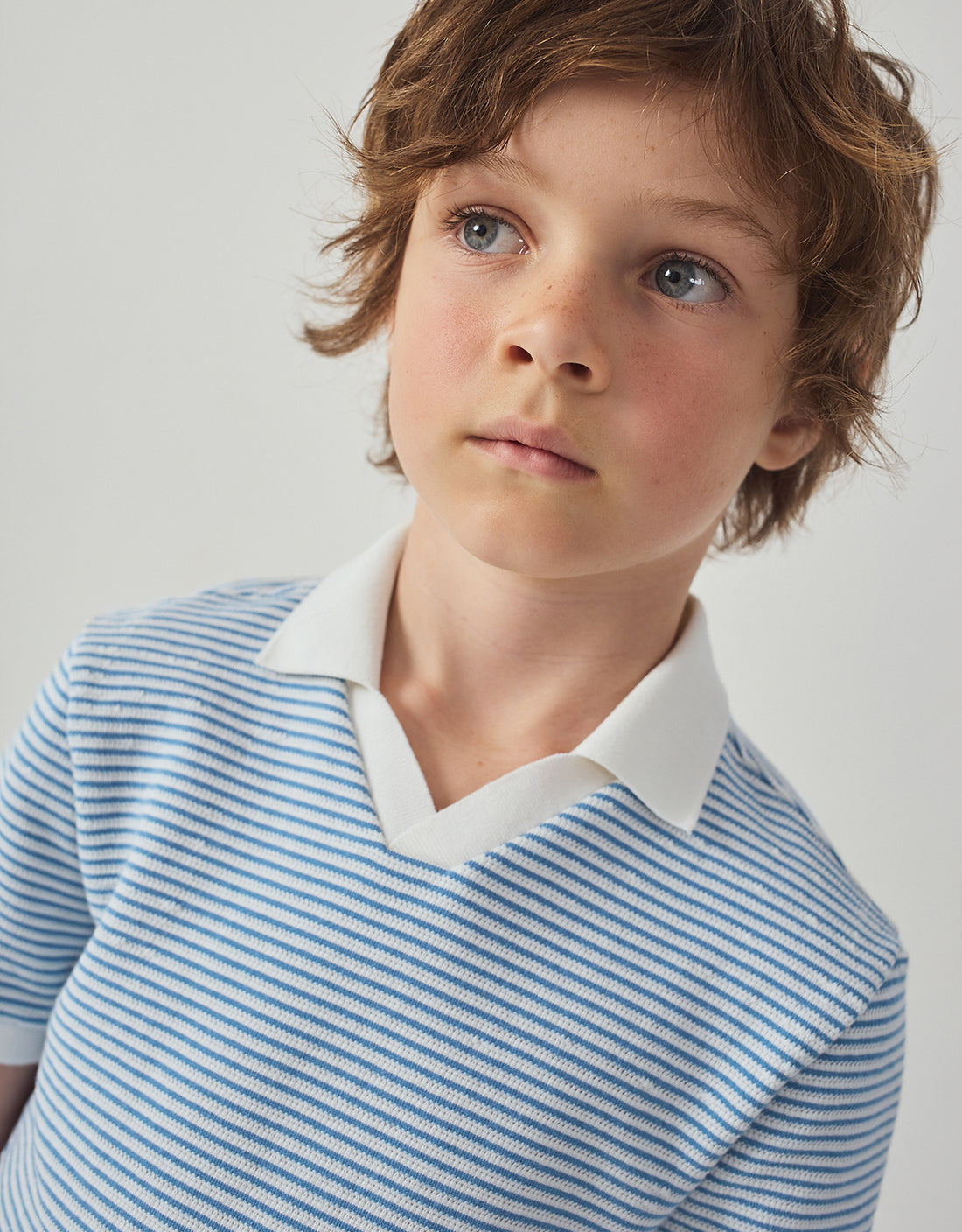 A young boy with light brown hair and blue eyes wears the Gingersnaps Boys' Husker Striped Terry Shirt, featuring blue and white stripes, as he looks slightly upward against a plain background.