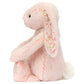 The Jellycat Blossom Blush Bunny ‘Cherry’ is a light pink plush toy with floral-patterned ears and feet, sitting upright with a fluffy tail and black eyes.