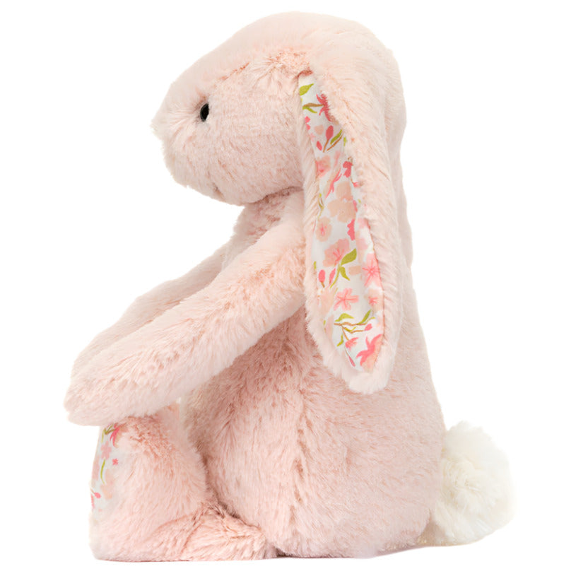 The Jellycat Blossom Blush Bunny ‘Cherry’ is a light pink plush toy with floral-patterned ears and feet, sitting upright with a fluffy tail and black eyes.