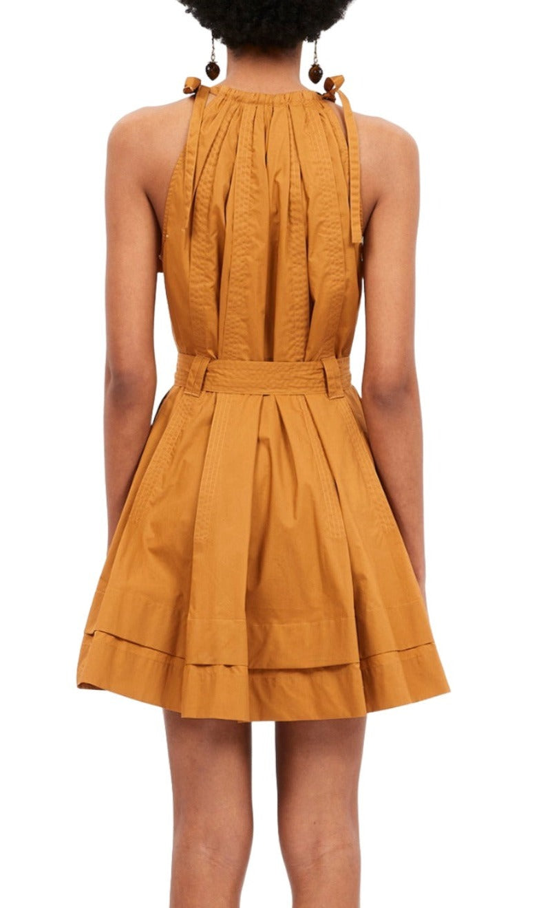 Woman wearing a sleeveless, mustard-colored Ulla Johnson Brienne Dress, Topaz crafted from cotton poplin, featuring gathered pleats and a flared skirt, viewed from the back. Adjustable strap ties add an extra touch of elegance to this stunning piece.