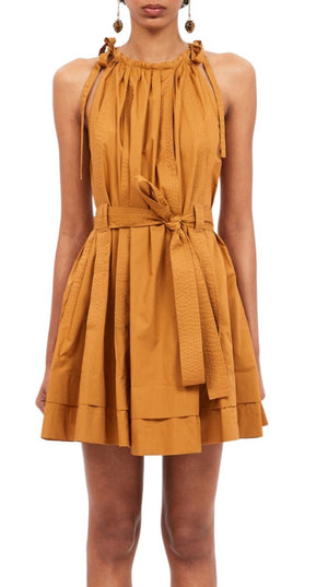 A person wearing the Ulla Johnson Brienne Dress, Topaz, a sleeveless, knee-length, burnt orange piece made from cotton poplin with a cinched waist and tiered skirt. The dress features adjustable strap ties at the halter neckline.