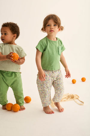 Two young children stand barefoot on a light background with oranges on the floor. One wears green pants and the Cozmo Baby Girls' Brisa Ruffle Pocket T-Shirt; the other wears floral pants and a lightweight cotton green shirt.