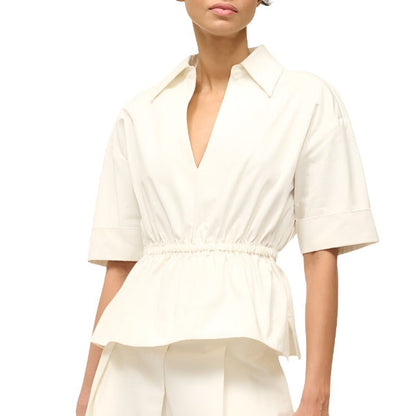 A person wearing the Staud Bristol Top, a cream-colored short-sleeve blouse with a v-neckline and an elastic waist, by Staud.