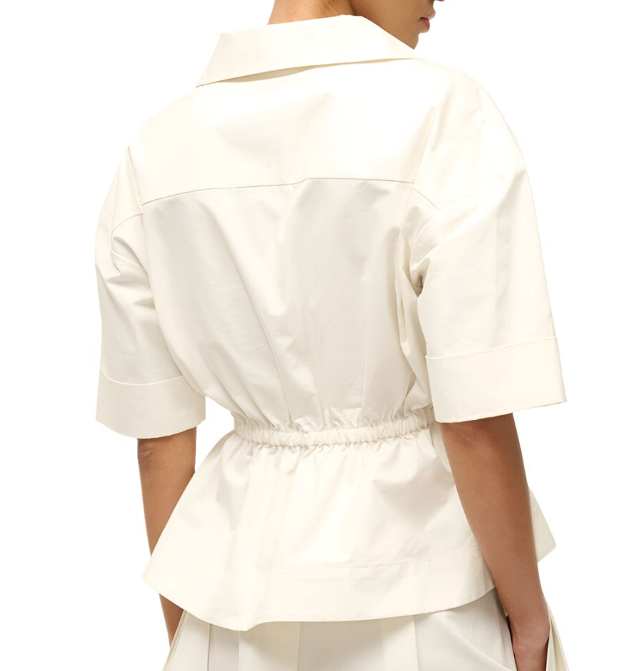 A person wearing the Staud Bristol Top, a white short-sleeved button-up peplum top with an elastic waist from the brand Staud, is shown from the back.