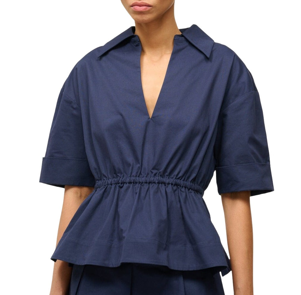 A person is wearing the Staud Bristol Top by Staud, a navy blue, short-sleeved, V-neck peplum top featuring a ruffled hem and cinched waist. The image shows the upper body only against a plain white background.