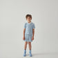A child stands against a plain white background, wearing a light blue collared shirt, blue socks, and Gingersnaps Boys' Dreyfus Striped Terry Drawstring Shorts.